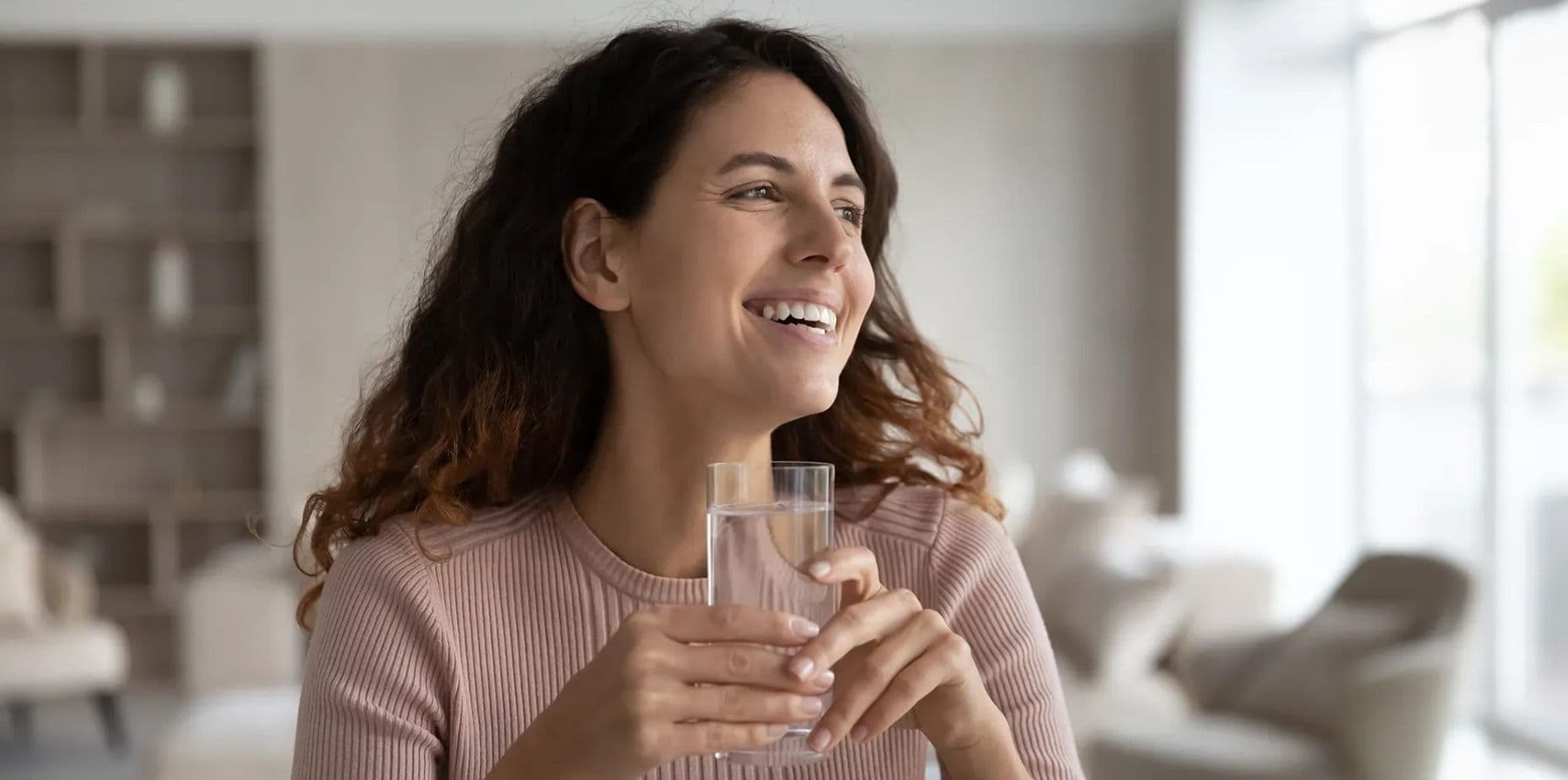 A woman with curly hair, wearing a pink sweater, smiles while holding a glass of water. She's in a bright, modern living room, conveying a sense of contentment.