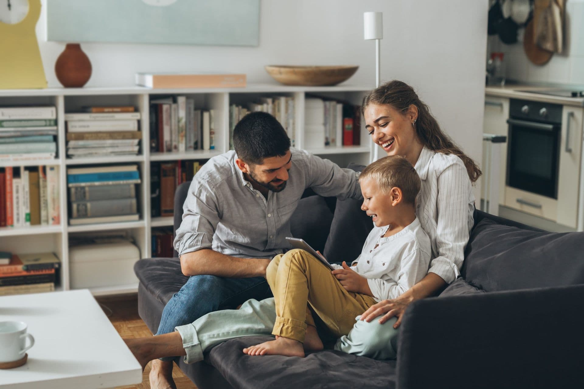 A happy family of three sits on a couch in a cozy living room, with bookshelves in the background. They are smiling and looking at a tablet together.