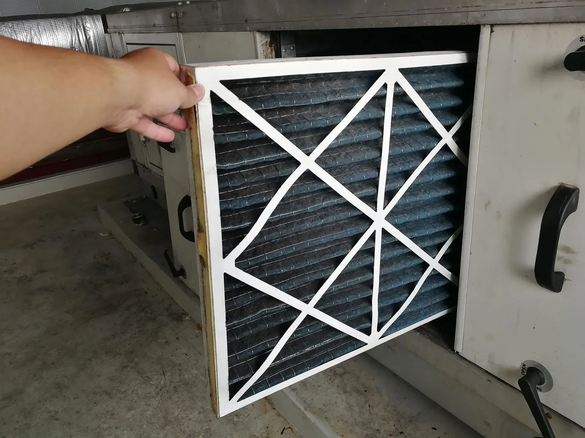 A hand holding a dusty air filter being removed from a large industrial HVAC unit, highlighting maintenance. The scene conveys functionality and upkeep.