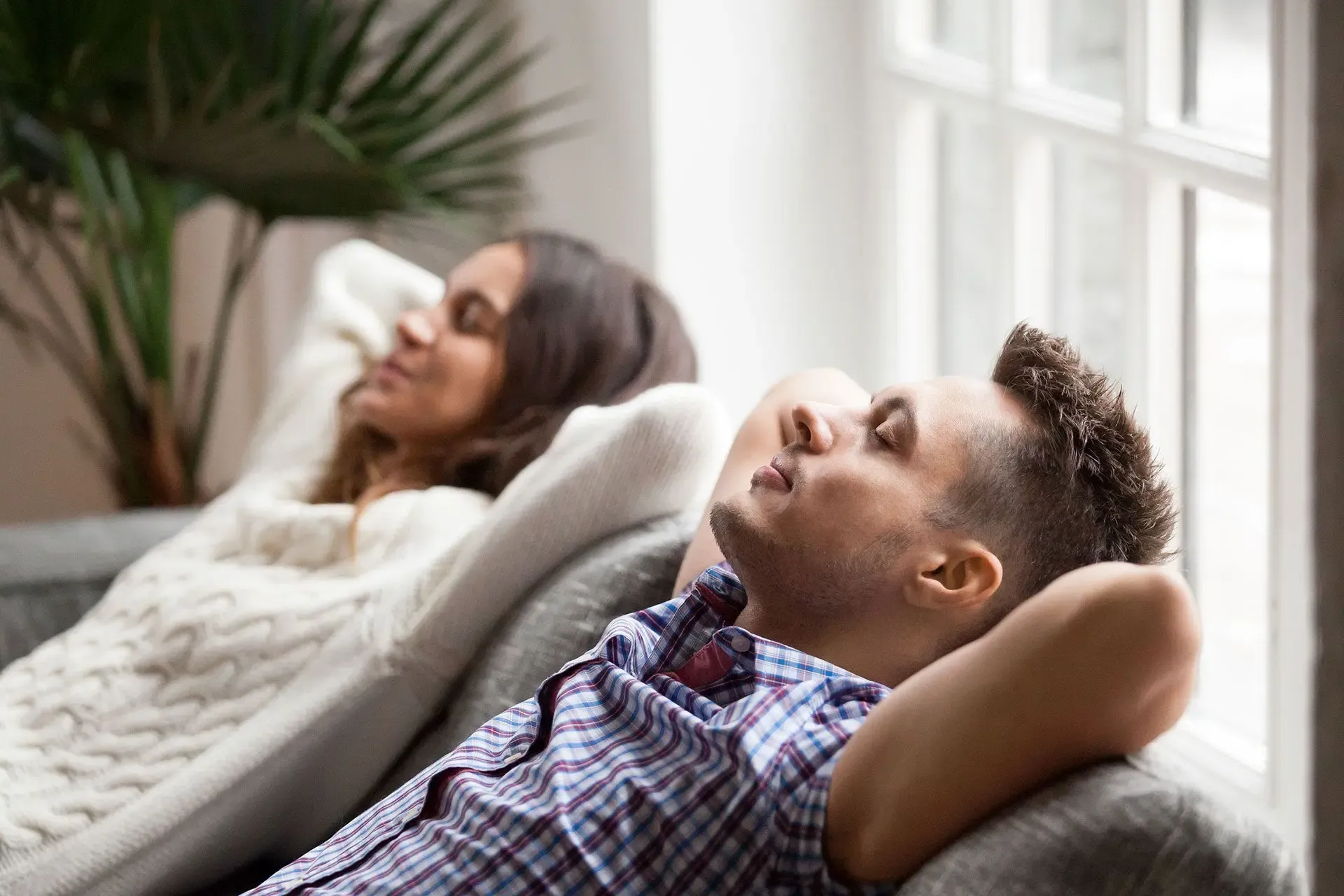 A man and woman relax on a couch with hands behind their heads, eyes closed, near a window with soft light. A plant is visible in the background.