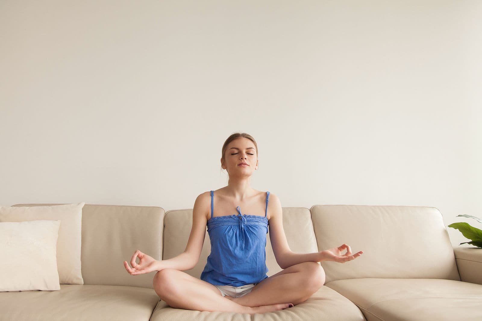 Woman in blue tank top sitting in lotus position on beige couch with eyes closed, meditating in calm, minimalist setting