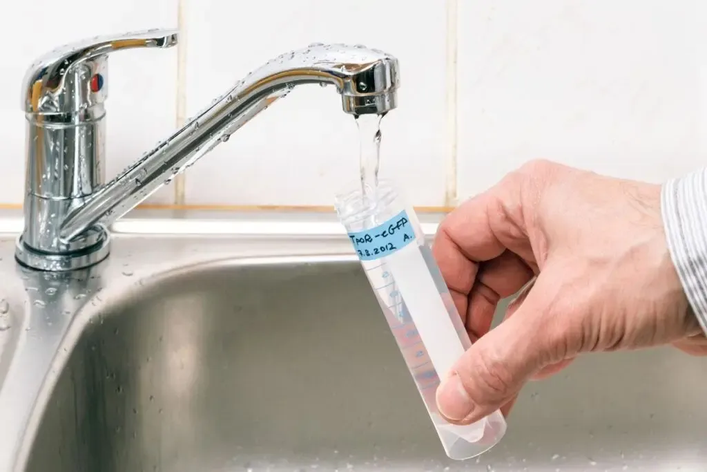 A hand holds a test tube labeled with text under a running kitchen faucet. Water flows into the tube, indicating a water collection or testing process.