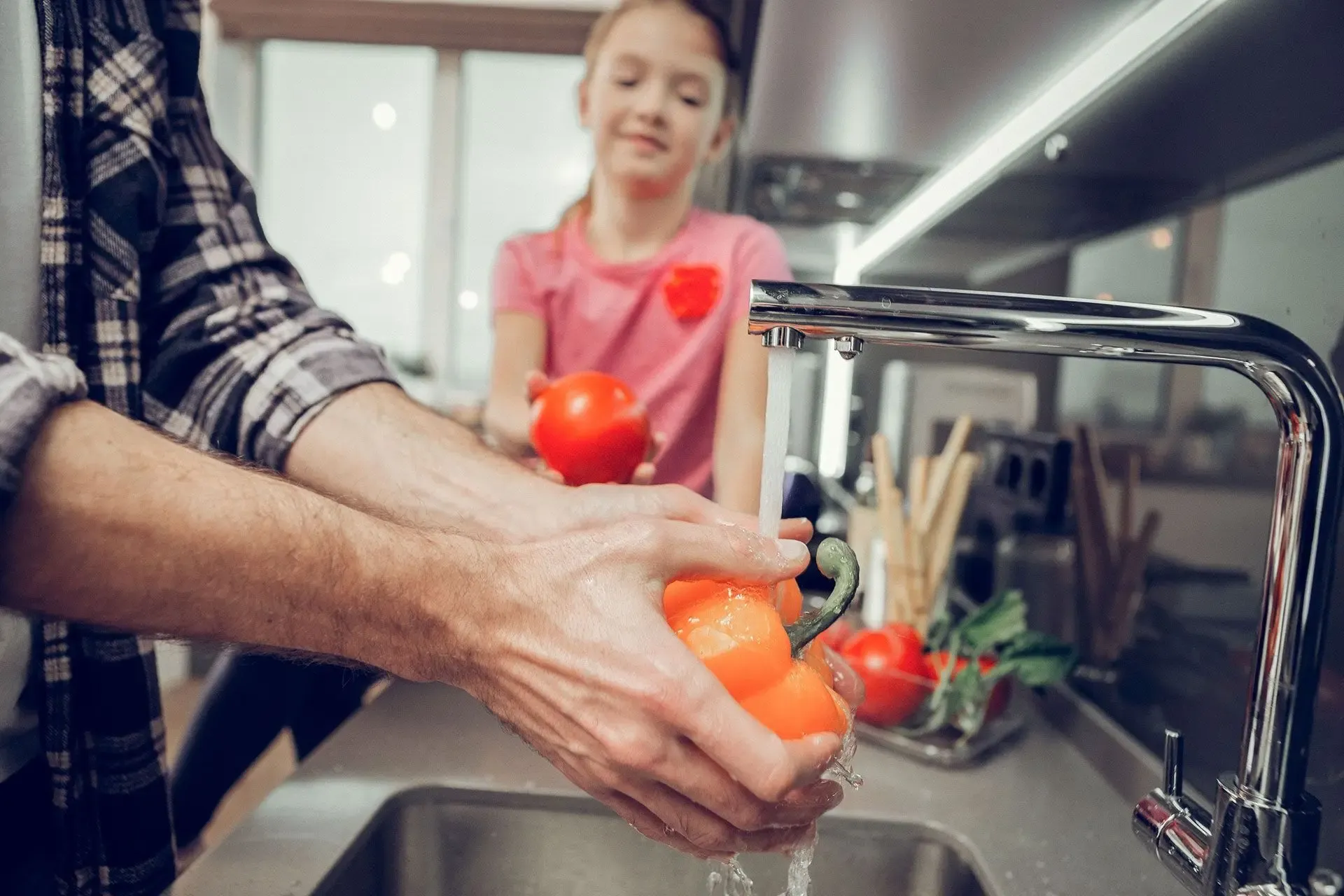 An adult washing tomatoes under a kitchen faucet with a child in a pink shirt holding a tomato, sharing a warm, collaborative kitchen moment.