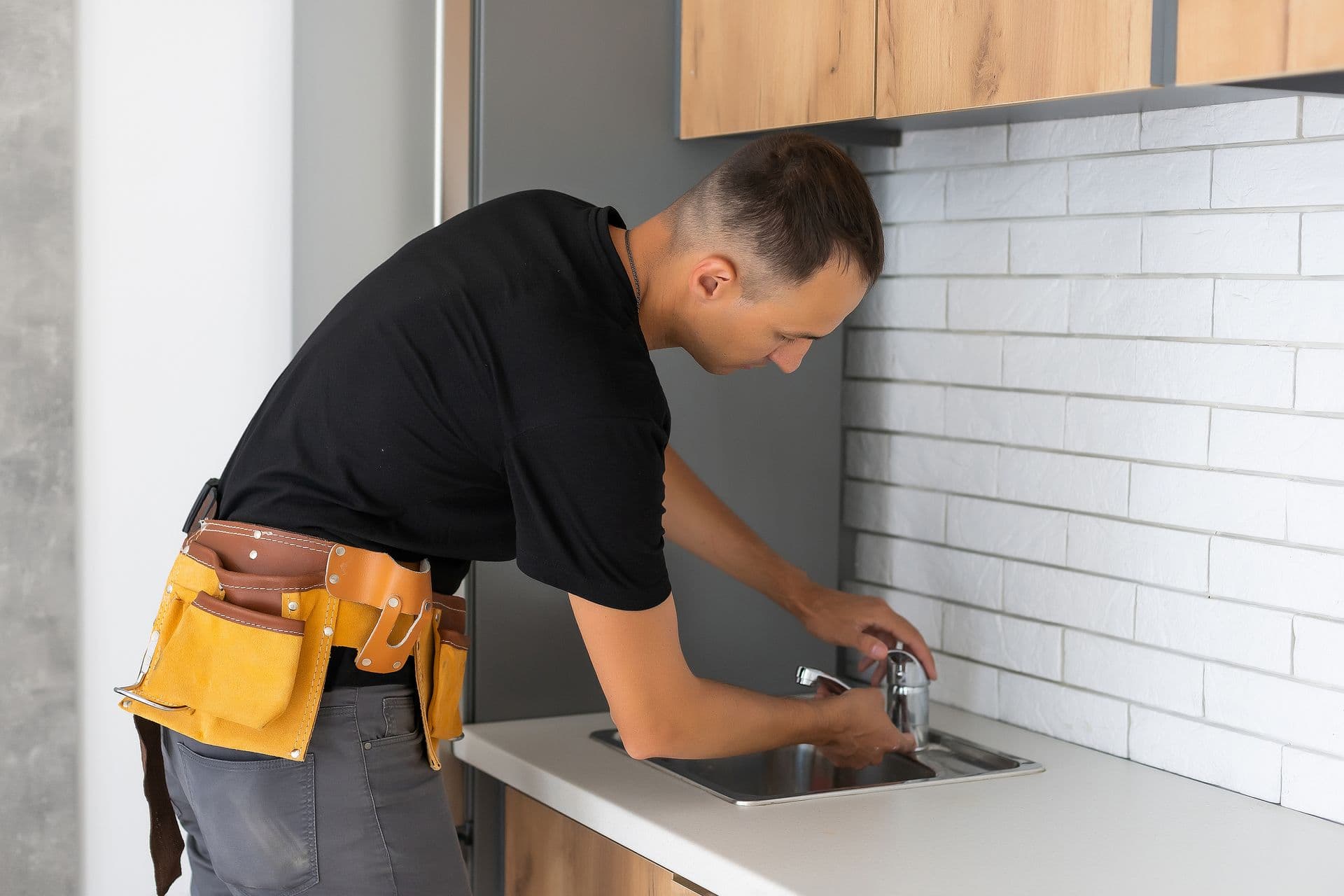 A man in a black shirt and work belt fixes a kitchen faucet, surrounded by modern cabinets and white brick backsplash, conveying focus and skill.