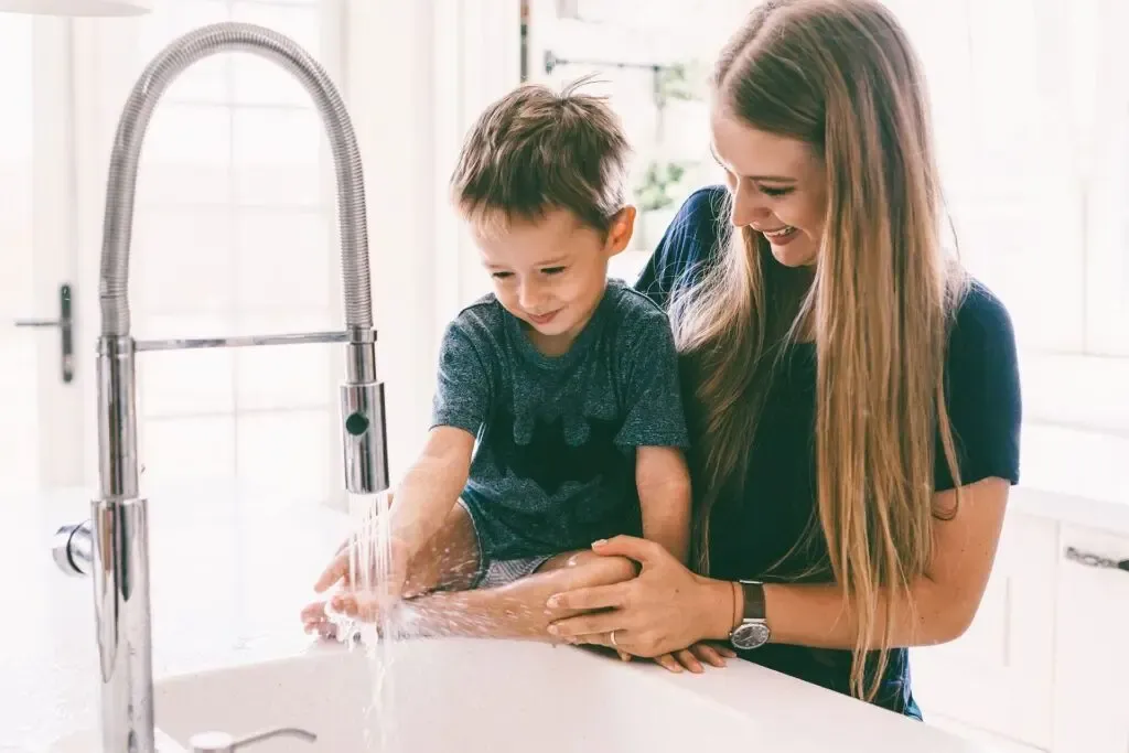 A woman and young child joyfully wash hands at a kitchen sink, sunlight streaming in. The scene conveys warmth and nurturing.