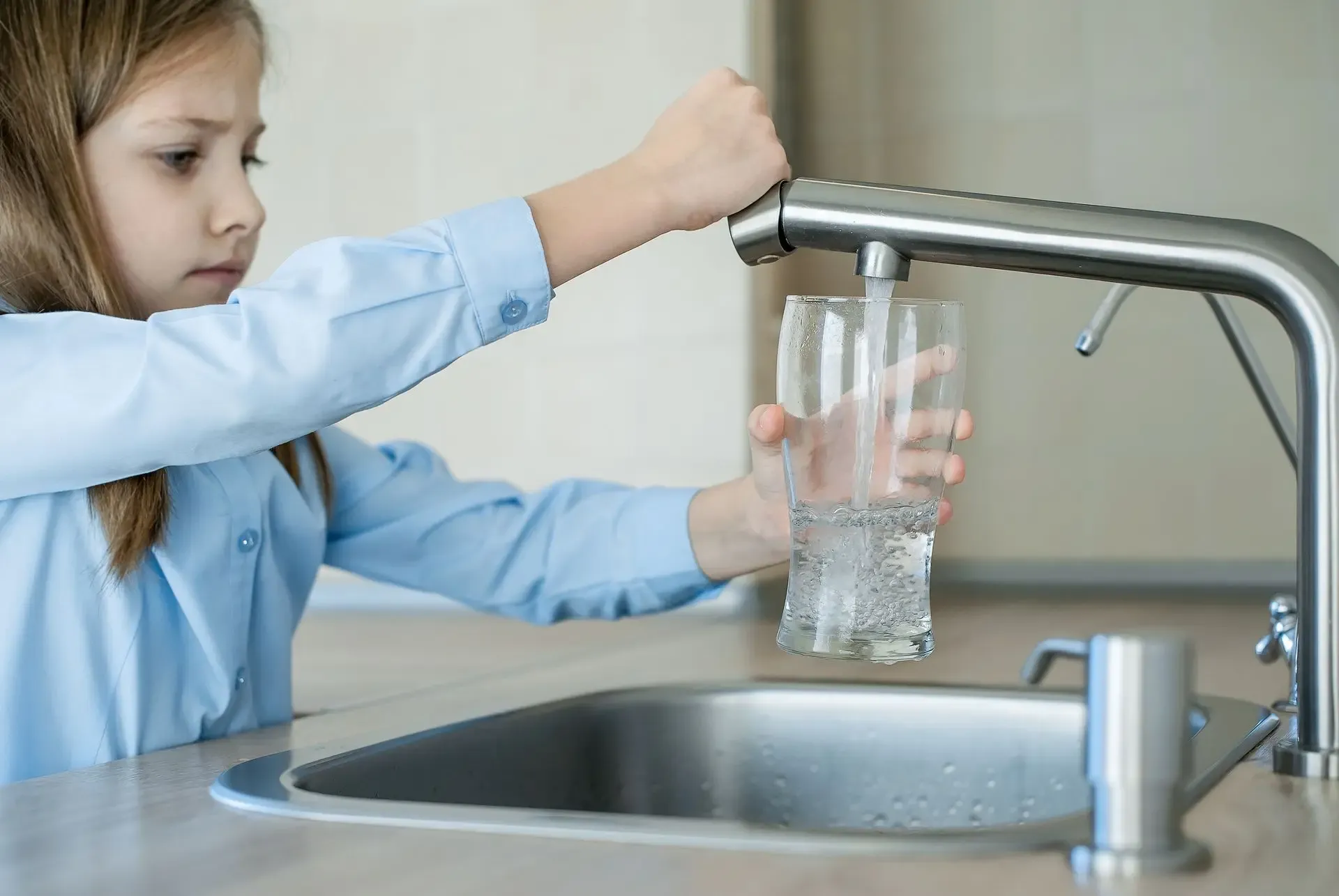 A young girl in a blue shirt fills a glass with water from a modern kitchen faucet. Her focused expression highlights careful attention to the task.