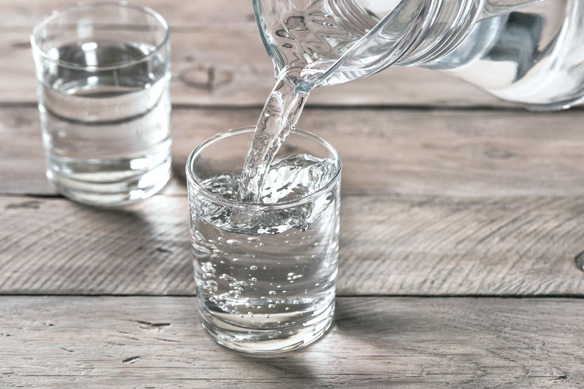 Water is being poured into two clear glasses placed on a wooden table.