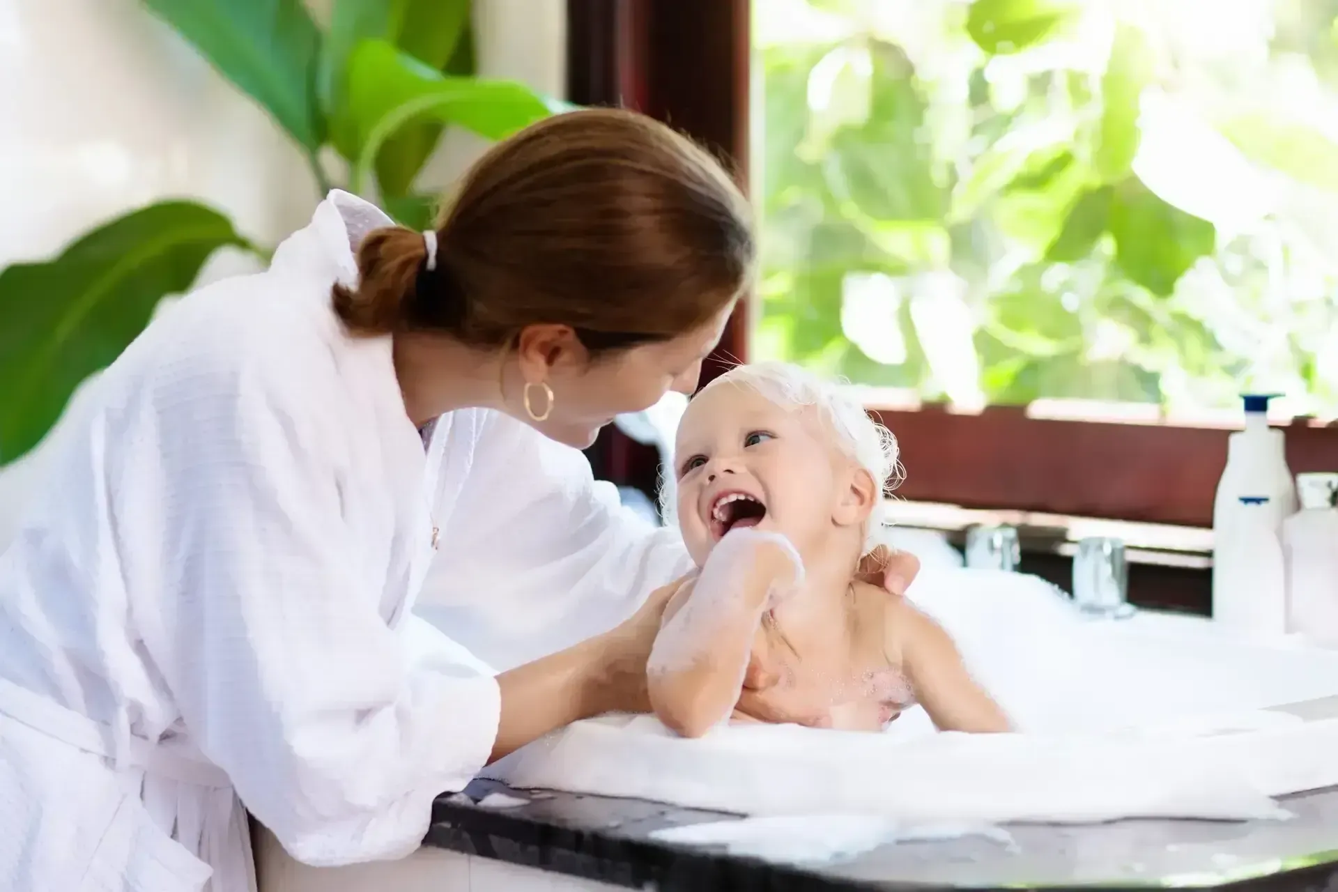 A woman in a white robe smiles at a laughing toddler in a bubble-filled bath. Bright natural light and green plants create a joyful, cozy atmosphere.