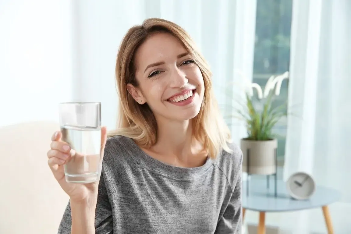Smiling woman holding a glass of water, wearing a gray shirt. She sits in a bright room with a plant and clock in the background, conveying a refreshing mood.