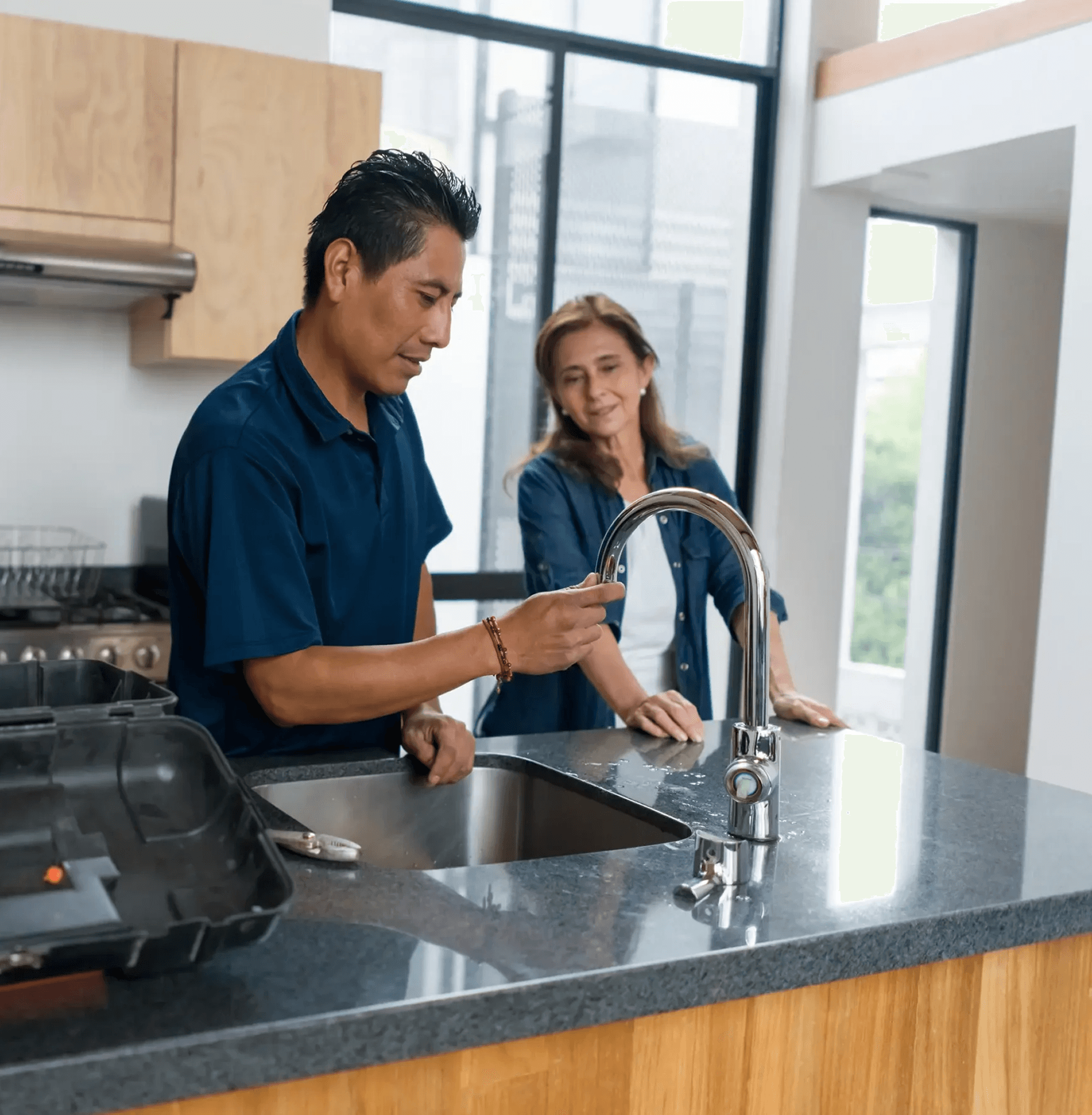 A man in a blue shirt checks a kitchen faucet while a woman watches. A toolbox is open on the counter. The scene conveys teamwork and focus.