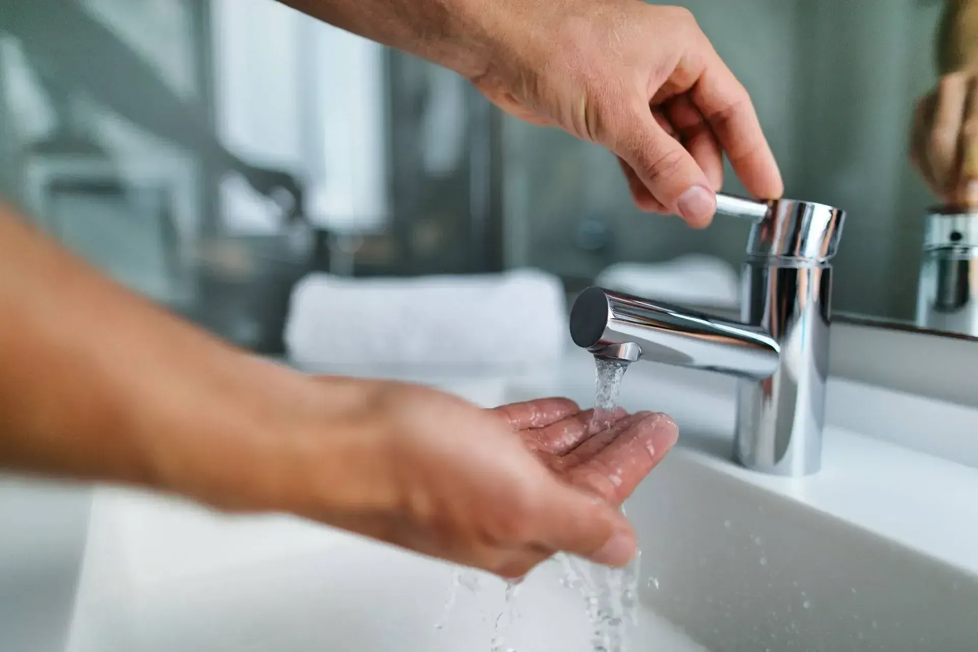 Hands under running water from a modern chrome faucet in a bathroom, evoking cleanliness and hygiene. A soft towel is blurred in the background.