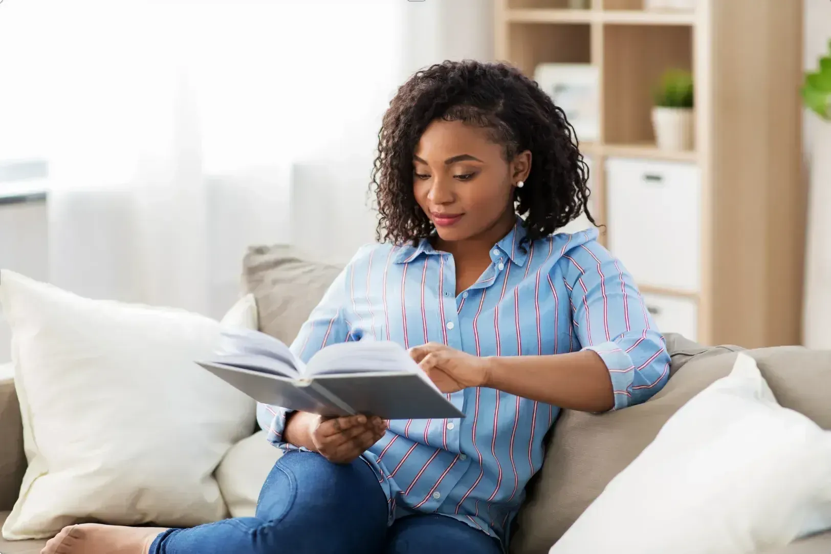 Woman in a blue striped shirt reading a book on a beige sofa, surrounded by white cushions. The room has a calm, cozy atmosphere.