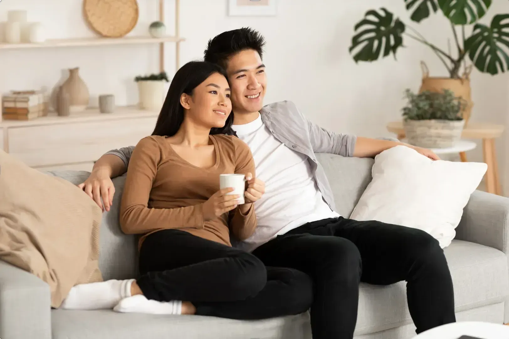A couple sits on a sofa in a cozy living room, smiling and relaxed. The woman holds a mug. The room is decorated with plants and neutral tones.