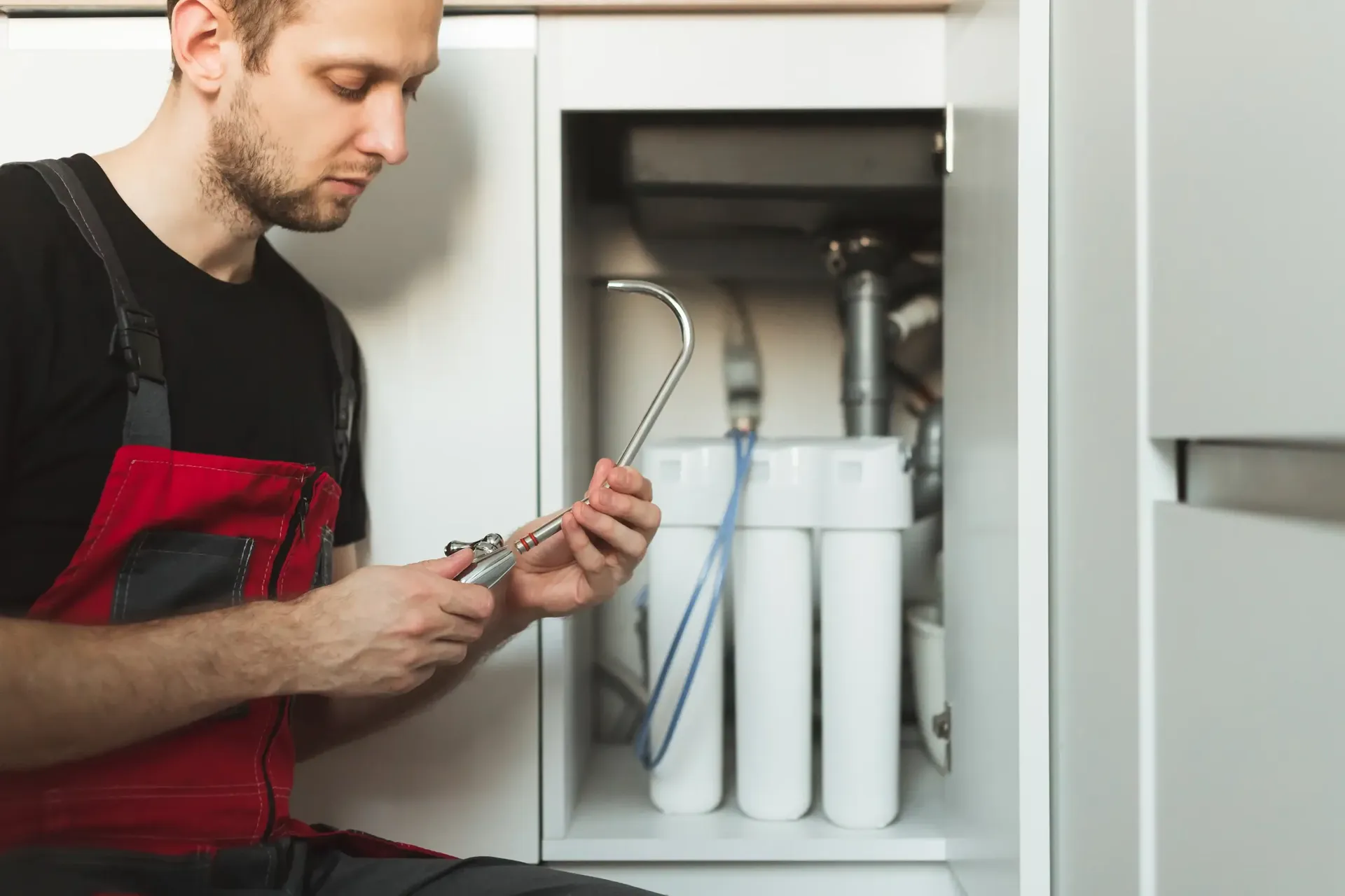 A plumber in red overalls examines a pipe tool beside an open cabinet with water filtration equipment, focused and intent on his repair work.