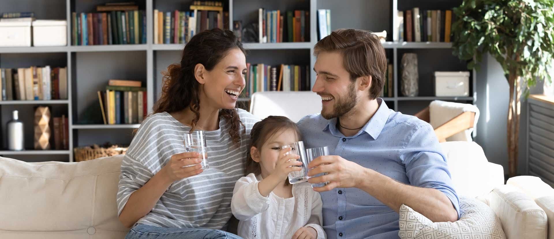 A happy family of three sits on a sofa, smiling and toasting with glasses of water. Behind them, shelves filled with books create a cozy background.